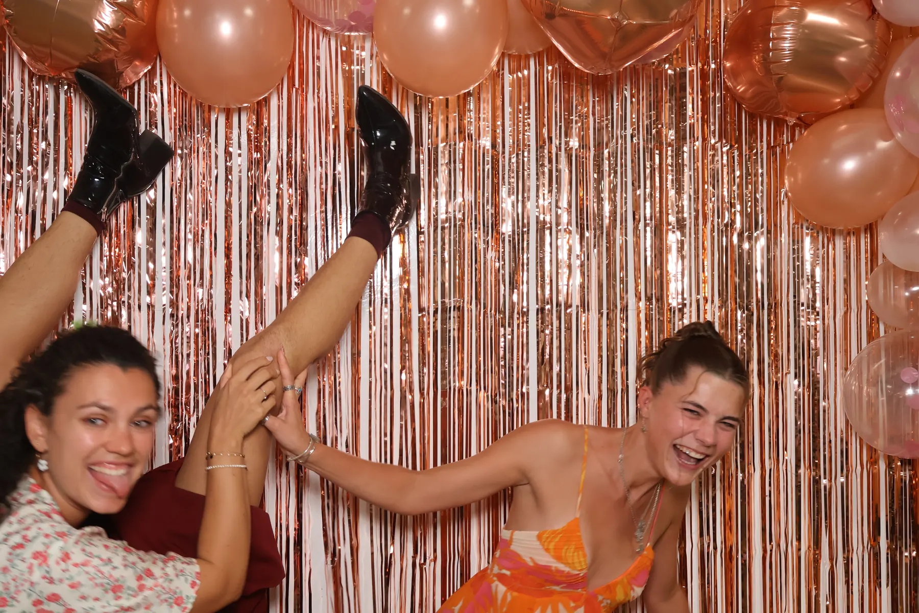 Deux invitées de mariage éclatent de rire, l'une levant la jambe, devant le rideau à franges cuivrées et les ballons du photobooth.