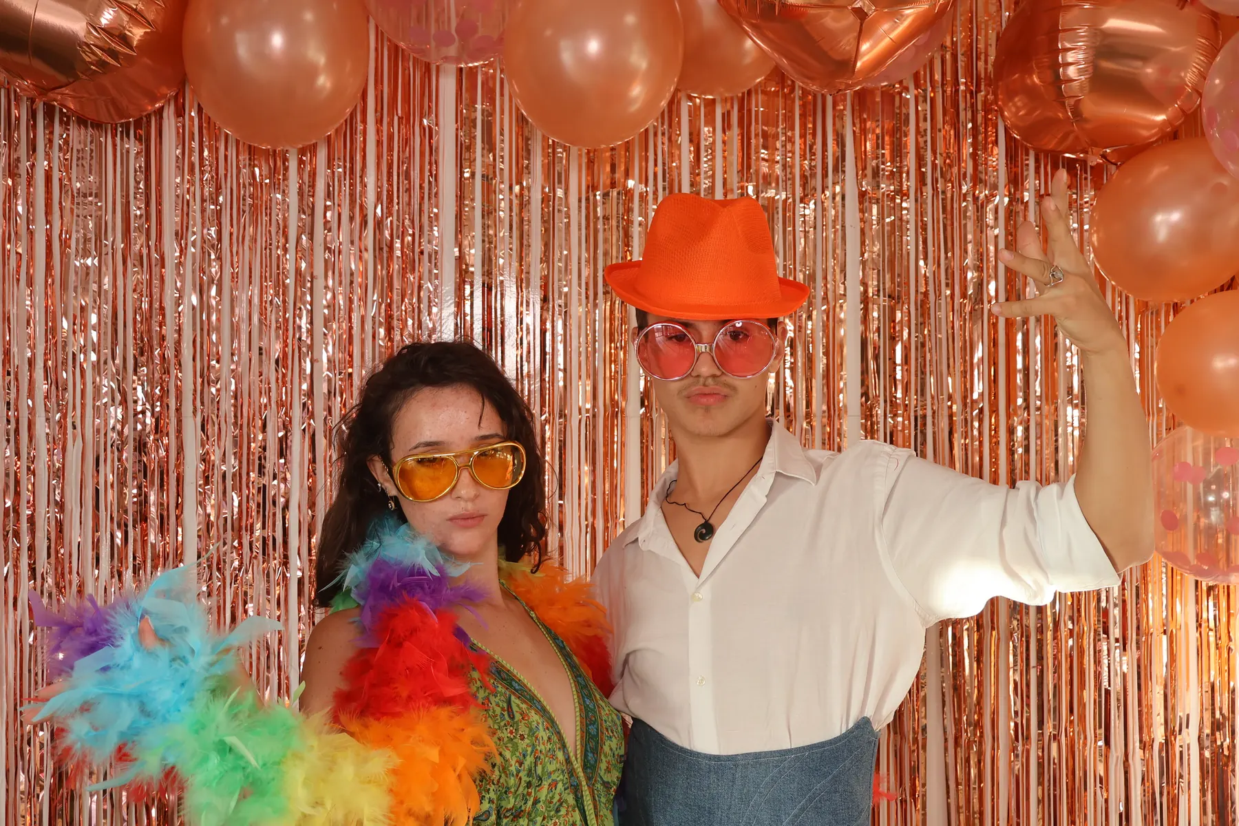 Un couple d'invités pose avec un boa multicolore, des lunettes jaunes et un chapeau orange devant le photobooth.