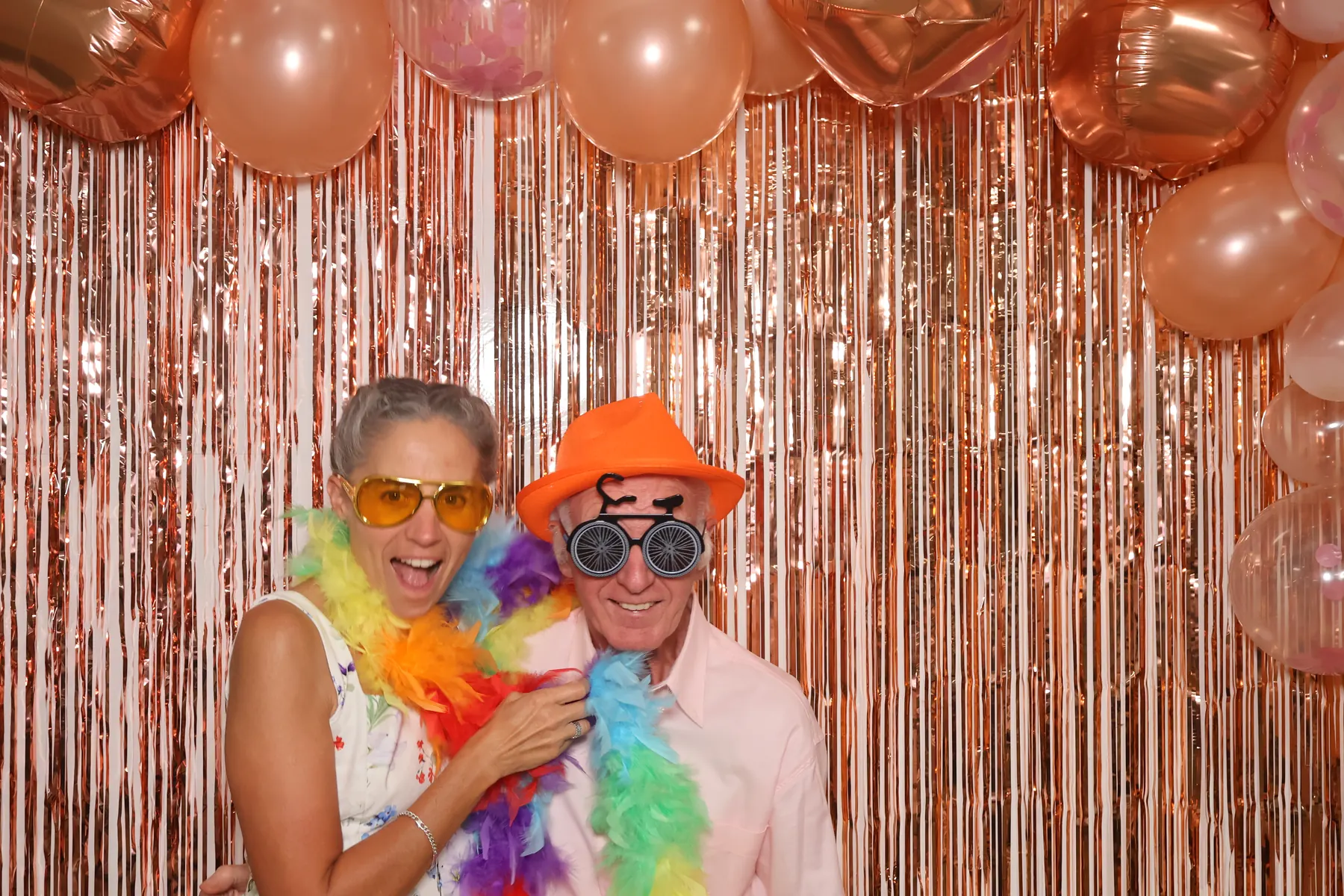 Un couple de seniors pose avec un chapeau orange, des lunettes fantaisie et des boas en plumes devant le photobooth, lors d'un mariage.