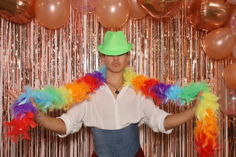 Un invité déploie un boa en plumes arc-en-ciel à bout de bras devant le rideau à franges cuivrées du photobooth, en plan large.