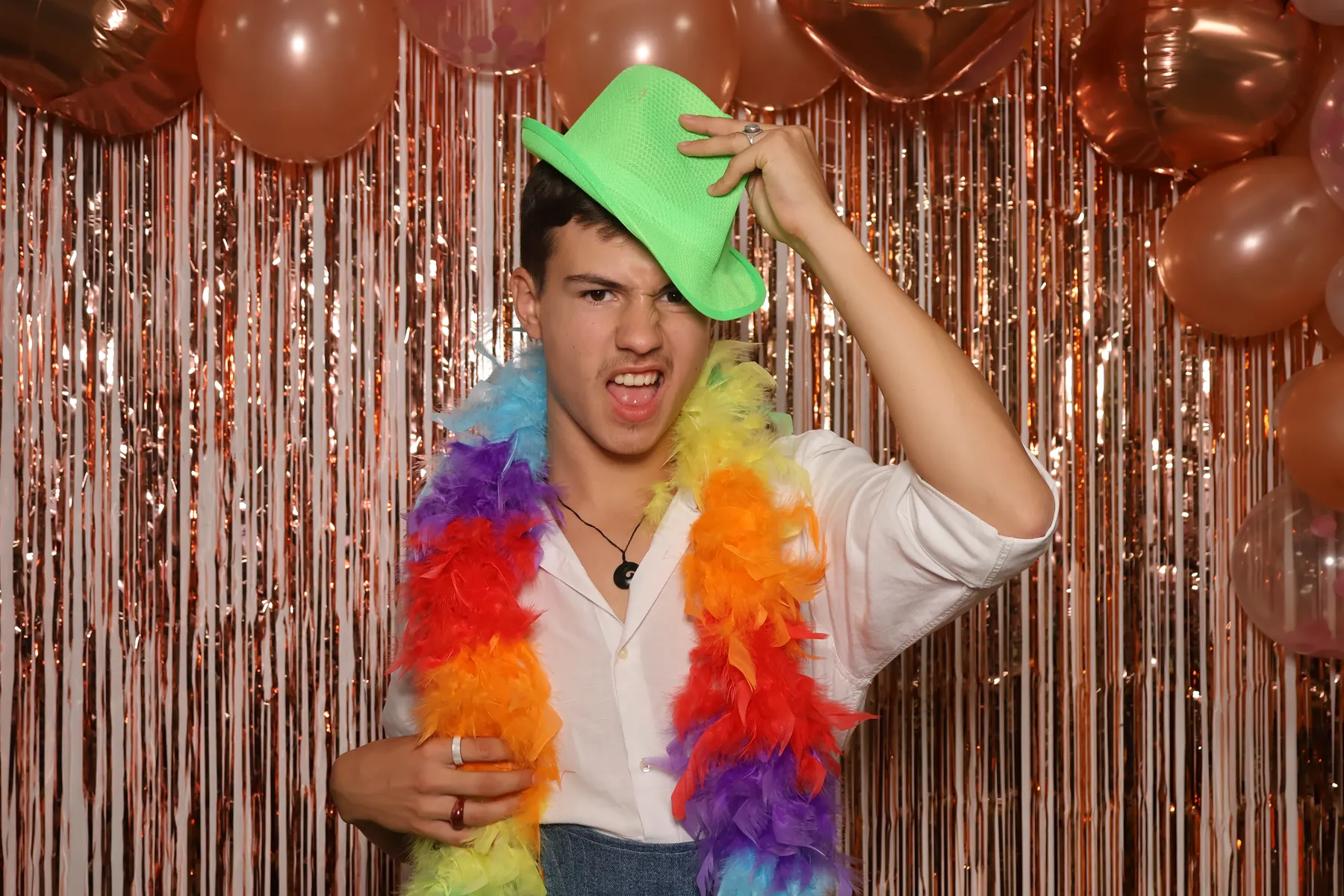 Un invité en chemise blanche ajuste un chapeau vert fluo, un boa en plumes autour du cou, devant le fond cuivre du photobooth.