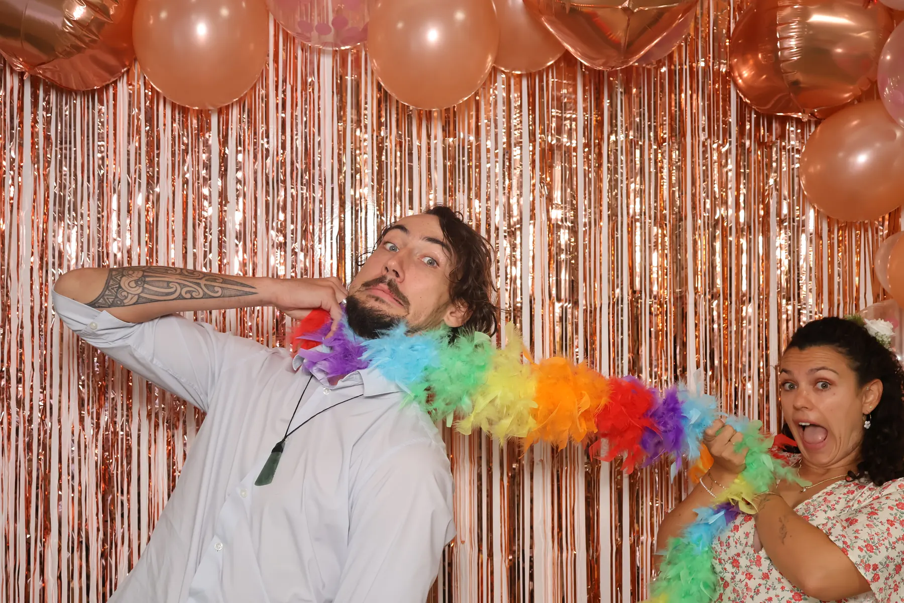 Deux invités de mariage tirent sur le même boa en plumes multicolores en riant, devant le rideau à franges cuivrées du photobooth.