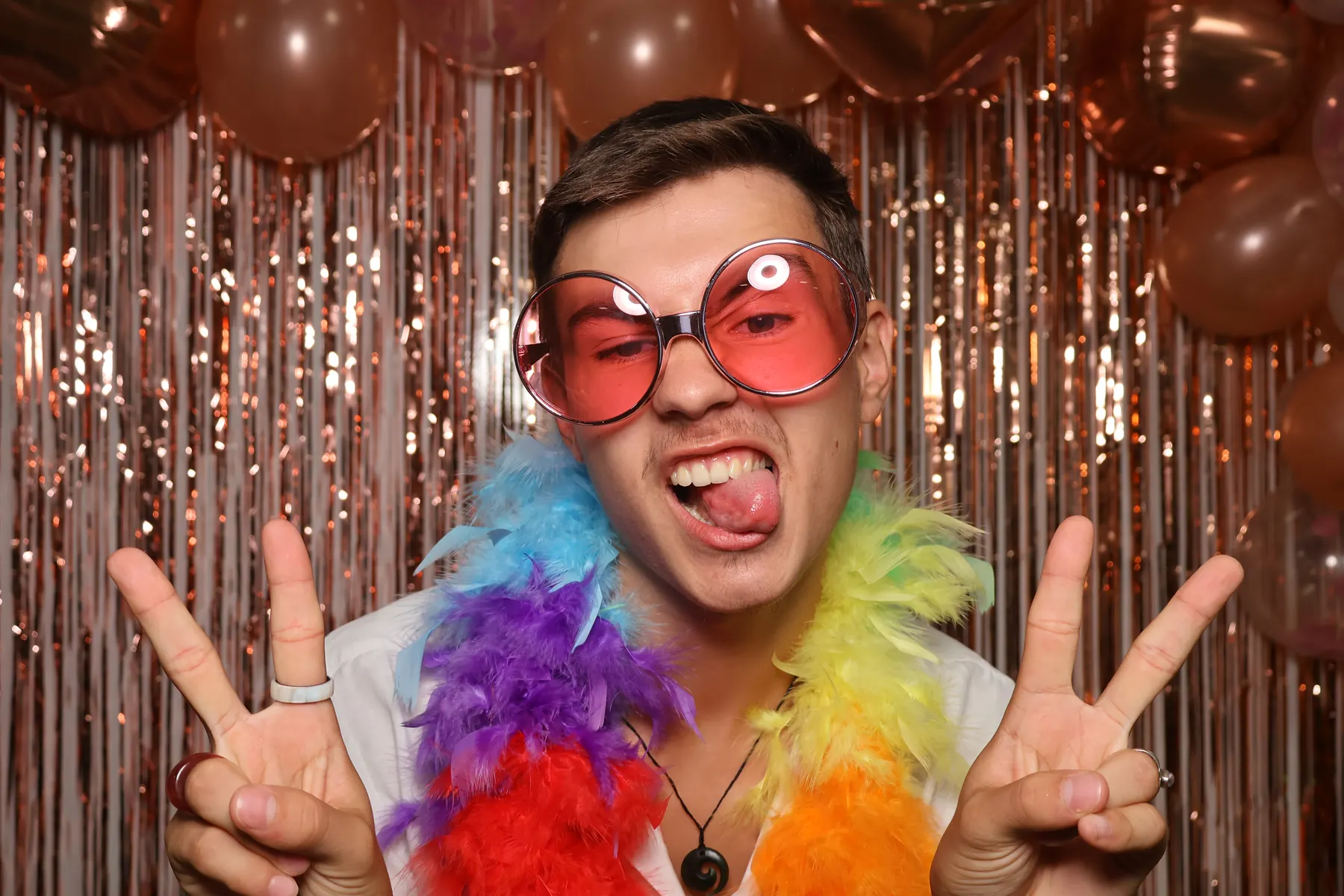 Portrait rapproché d'un invité qui tire la langue derrière des lunettes roses géantes, doigts en V, devant le fond cuivre du photobooth.