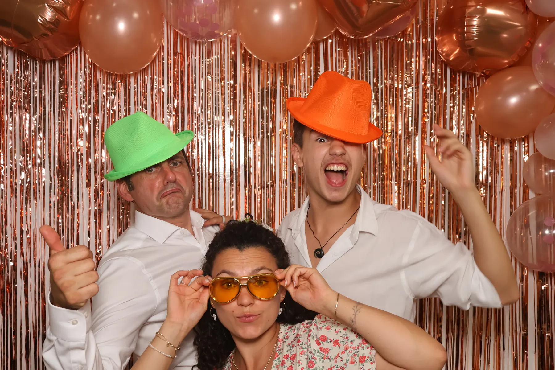Trois invités posent avec des chapeaux vert et orange et des lunettes jaunes devant le rideau à franges du photobooth.