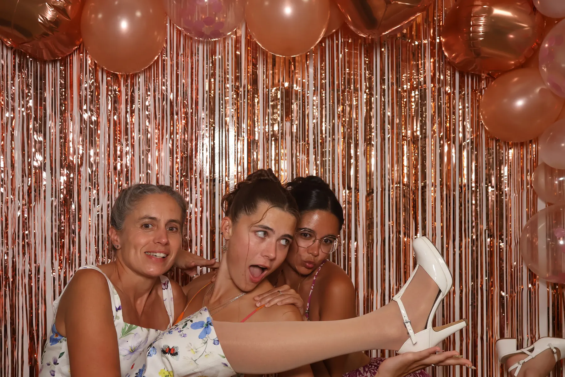 Trois invitées se serrent en riant devant le fond à franges cuivrées du photobooth, une chaussure à talon brandie au premier plan.