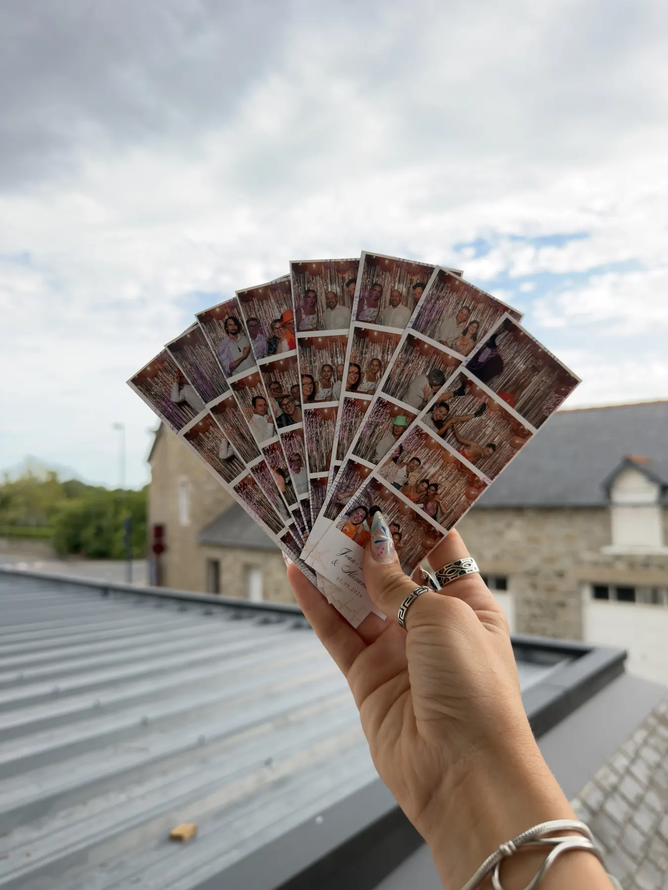 Une main tient en éventail une dizaine de tirages photobooth d'un mariage, devant des toits d'ardoise et un ciel nuageux de Bretagne.