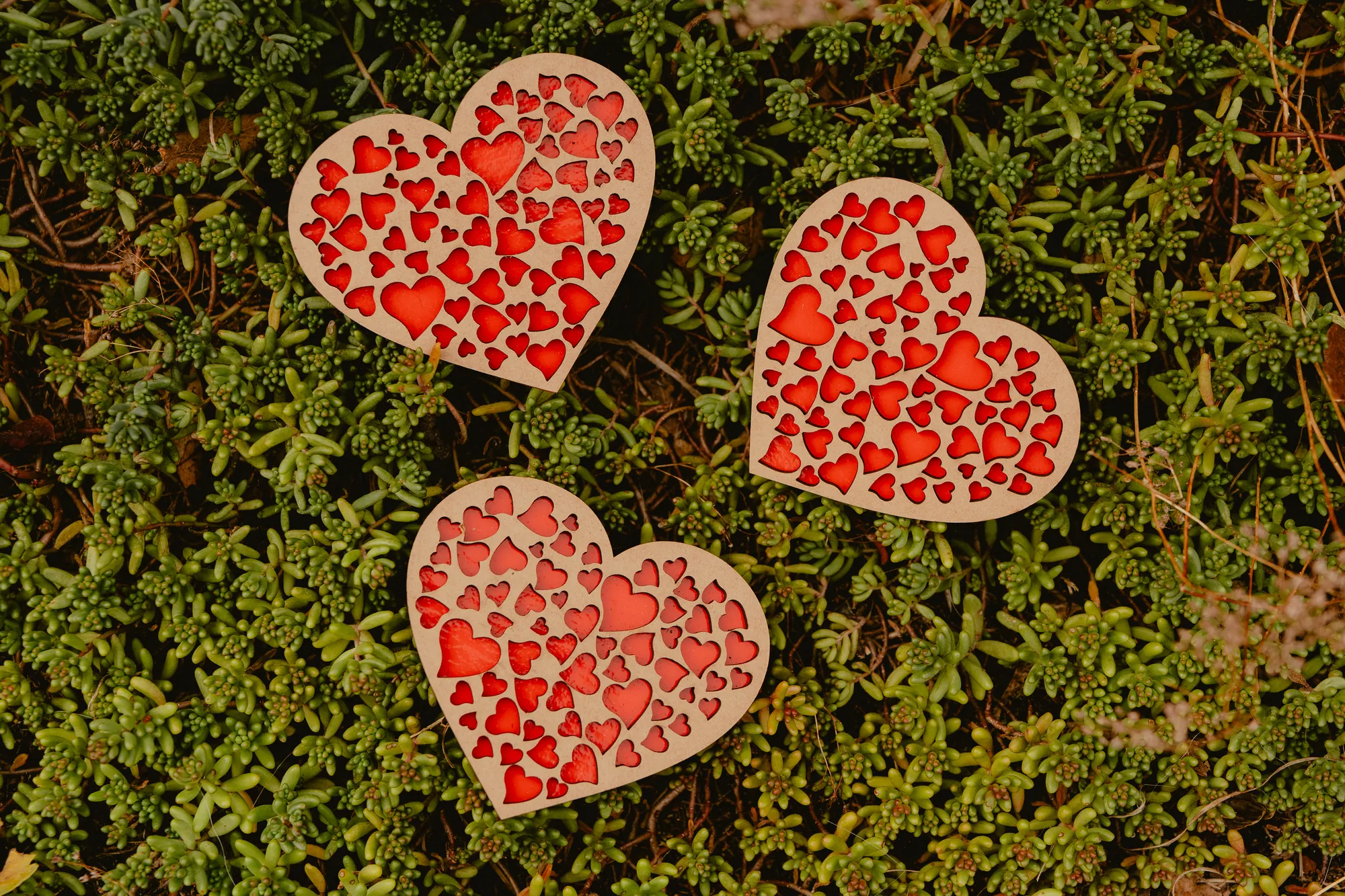 Trois magnets en bois en forme de cœur, découpés de petits cœurs sur fond rouge.